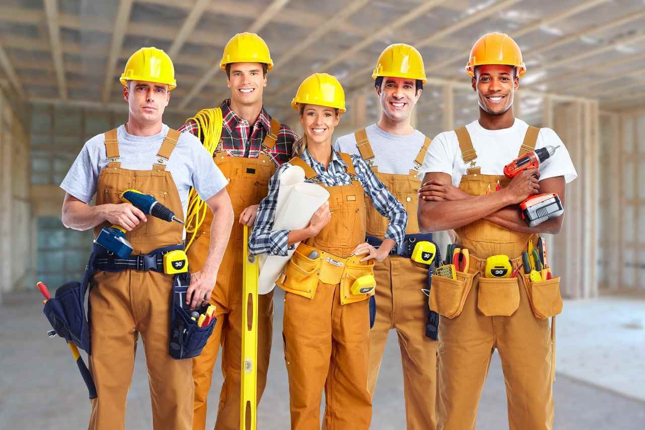 team of five electricians in hardhats and coveralls smiling at camera with new construction site in background