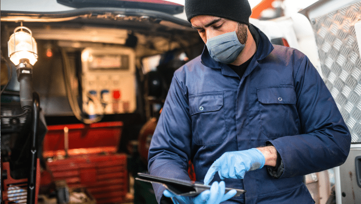 A technician on a field service call using his tablet while standing by his truck, he is wearing a face mask and gloves.