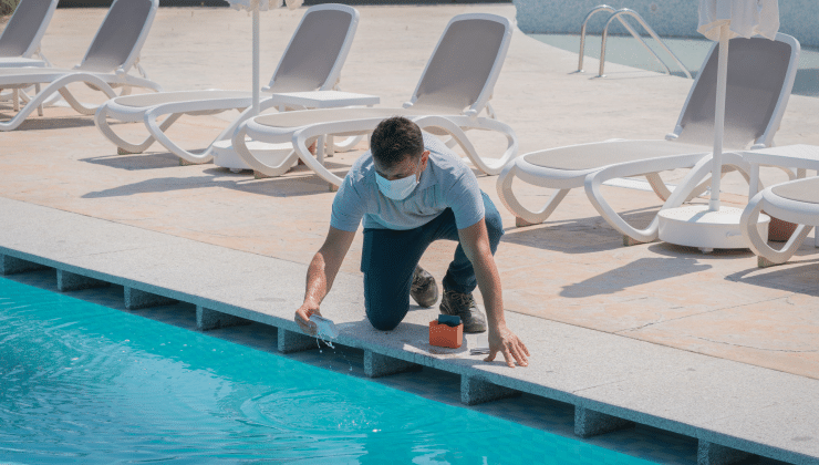 A male pool service technician kneels by a pool testing pH levels in the water